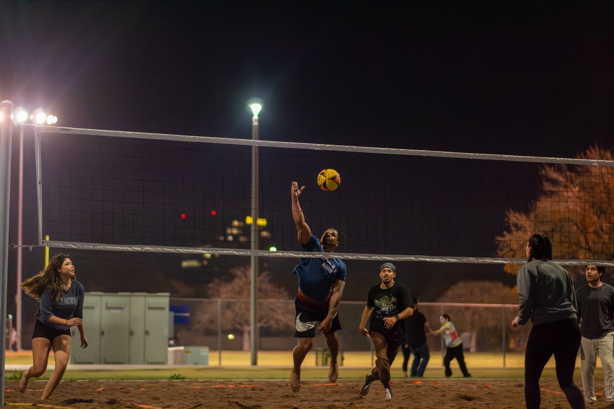 Adults mid-rally at a night sand-volleyball game on a floodlit outdoor court