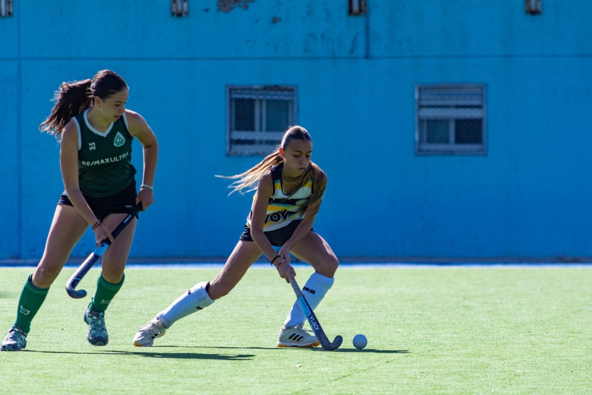 Two young field hockey players, one in a green kit and one in yellow and black, chase the ball stick-to-stick in a tight dribbling duel on a turf pitch, a weathered blue wall behind them.